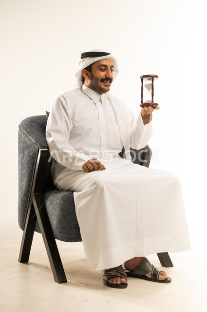مراقبة مرور الوقت بايماءات السعادة والسرور، إدراك قيمة اللحظات الثمينة في الحياة، التخطيط لتحقيق الأهداف في المستقبل، انتظار شيء ما بفارغ الصبر، بورتريه لرجل عربي خليجي قطري يرتدي الثوب التقليدي والغترة يجلس على كرسي ويمسك الساعة الرملية، خلفية بيضاء