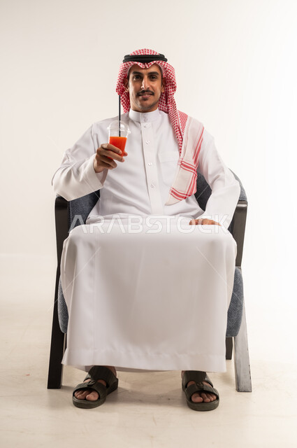 شرب المشروبات الغازية المنكهة اللذيذة، الحصول على عصير بارد ومنعش، الاستمتاع بتناول المشروب المفضل، بورتريه لشاب عربي خليجي بحريني يرتدي الثوب التقليدي والشماغ يجلس على كرسي يمسك بيده كأس من كوكتيل الفواكه الطازج، الطول الكامل للجسم، خلفية بيضاء