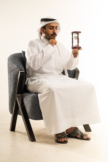 Planning to achieve goals in the future, waiting for something impatiently, portrait of a Qatari Gulf Arab man wearing traditional thobe and ghutra sitting on a chair and holding an hourglass, watching the passage of time with gestures of seriousness and integration, realizing the value of precious moments in life, white background
