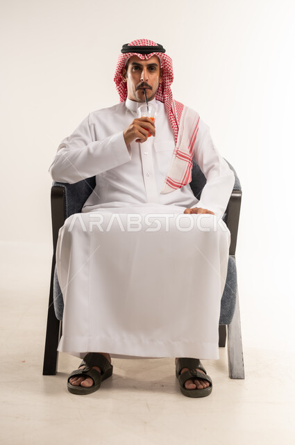 الحصول على عصير بارد ومنعش، الاستمتاع بتناول المشروب المفضل، شرب المشروبات الغازية المنكهة اللذيذة، بورتريه لشاب عربي خليجي بحريني يرتدي الثوب التقليدي والشماغ يجلس على كرسي يمسك بيده كأس من كوكتيل الفواكه الطازج، الطول الكامل للجسم، خلفية بيضاء