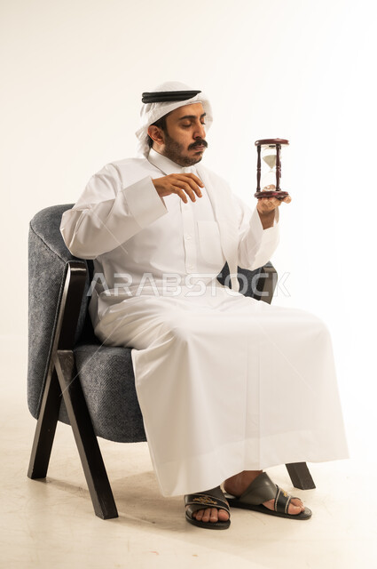 Realizing the value of precious moments in life, planning to achieve goals in the future, waiting for something impatiently, portrait of a Qatari Gulf Arab man wearing traditional thobe and ghutra sitting on a chair and holding an hourglass, watching the passage of time with gestures of seriousness and integration, white background