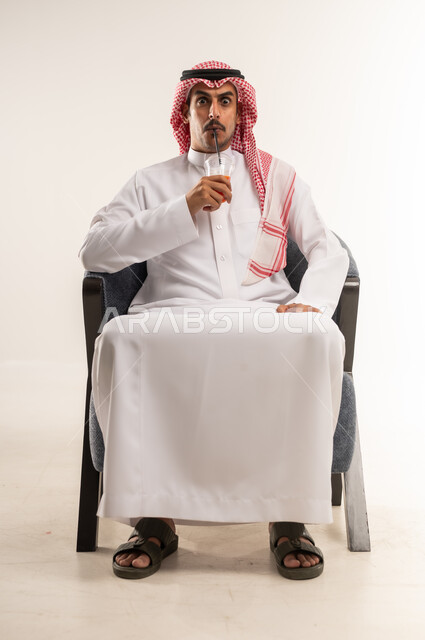 Enjoying a favorite drink, having a cold and refreshing juice, drinking delicious flavored soft drinks, portrait of a young Arab Gulf Bahraini man wearing a traditional thobe and shemagh sitting on a chair holding a glass of fresh fruit cocktail in his hand, full body length, white background
