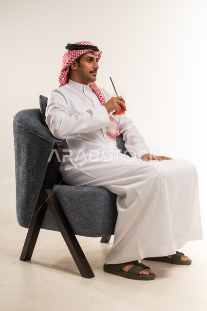 الحصول على كأس من العصير البارد والمنعش، الجلوس ومحاولة الراحة والاستمتاع بأوقات الفراغ، بورتريه من الجانب لشاب عربي خليجي بحريني يرتدي الثوب التقليدي والشماغ يجلس على كرسي ويمسك بيده كوب من المشروب الصيفي المفضل، الطول الكامل للجسم، خلفية بيضاء