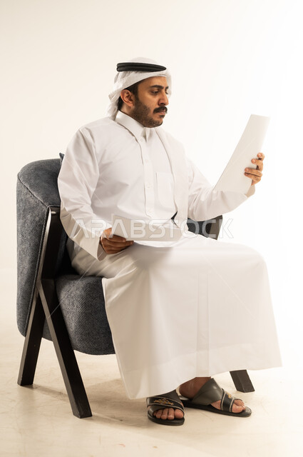 متابعة الخطط والمشاريع، قراءة الملاحظات والمعلومات، بورتريه لرجل عربي خليجي قطري يرتدي الثوب التقليدي والغترة يجلس على كرسي مريح يقوم بتدقيق الاوراق بتعابير التركيز والتمعن، مفهوم انجاز العمل وتحقيق المهام المطلوبة، خلفية بيضاء