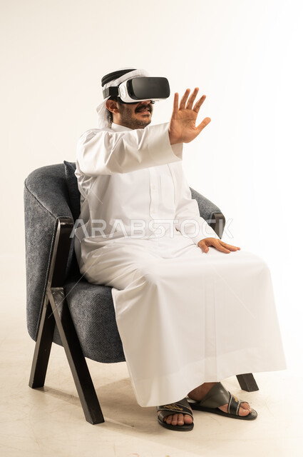 المتعة والتسلية بالمشاهدة عبر نظارة الواقع الافتراضي, بورتريه لرجل عربي خليجي قطري يرتدي الثوب التقليدي والغترة يجلس على اريكة مريحة يضع نظارات ثلاثية الأبعاد يقوم برفع  كف يده للأعلى، تعابير الاندماج والتوقف عن فعل امر ما, خلفية بيضاء