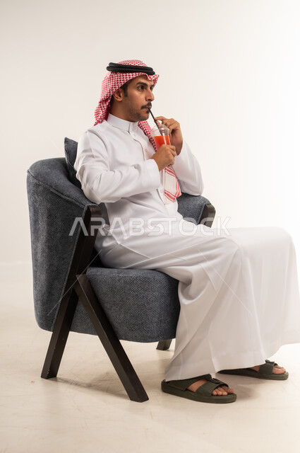 تناول المشروب المفضل والاستمتاع بمشاهدة الأفلام، شرب المشروبات الغازية المنكهة اللذيذة، الحصول على عصير بارد ومنعش، بورتريه من الجانب لشاب عربي خليجي بحريني يرتدي الثوب التقليدي والشماغ يجلس على كرسي ويمسك بيده كأس من الكوكتيل، الطول الكامل للجسم، خلفية بيضاء