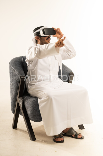 الاشارة الى مكان محدد بتعابير الاندماج, محاولة النقر على شيء ما, التسلية بالمشاهدة عبر نظارة الواقع الافتراضي, بورتريه لرجل عربي خليجي قطري يرتدي الثوب التقليدي والغترة يجلس على اريكة مريحة يضع نظارات ثلاثية الأبعاد يقوم برفع اصبع السبابة للاعلى، خلفية بيضاء