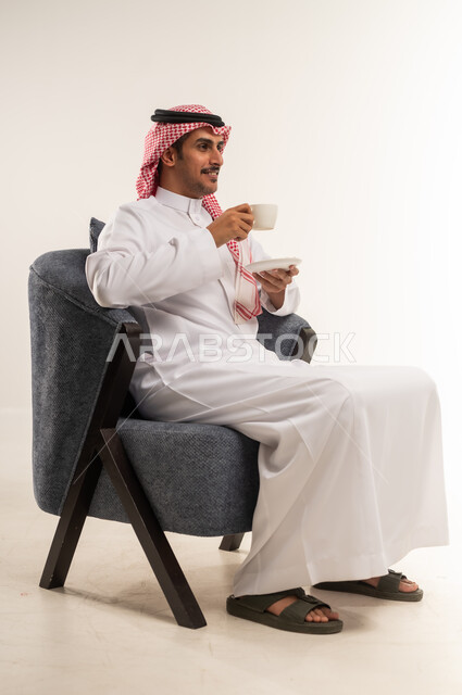 محاولة الحصول على القليل من الراحة، الاسترخاء وتناول مشروبات دافئة، الاستمتاع بكوب من القهوة الساخنة الغنية، بورتريه من الجانب لشاب عربي خليجي بحريني يرتدي الثوب التقليدي والشماغ يجلس على كرسي ويمسك بيده فنجان من مشروبه المفضل، الطول الكامل للجسم، خلفية بيضاء
