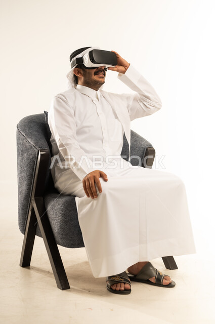 التفاعل والاندماج ومحاكاة العالم الخيالي، تعابير السعادة والسرور، المتعة والتسلية بالمشاهدة عبر نظارة الواقع الافتراضي, بورتريه لرجل عربي خليجي قطري يرتدي الثوب التقليدي والغترة يجلس على اريكة مريحة يضع نظارات ثلاثية الأبعاد، خلفية بيضاء