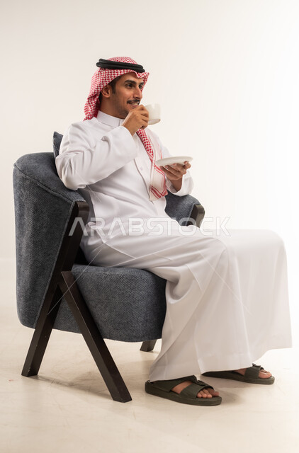 الاستمتاع بكوب من القهوة الساخنة الغنية، محاولة الحصول على القليل من الراحة، الاسترخاء وتناول مشروبات دافئة، بورتريه من الجانب لشاب عربي خليجي بحريني يرتدي الثوب التقليدي والشماغ يجلس على كرسي ويمسك بيده فنجان من مشروبه المفضل، الطول الكامل للجسم، خلفية بيضاء