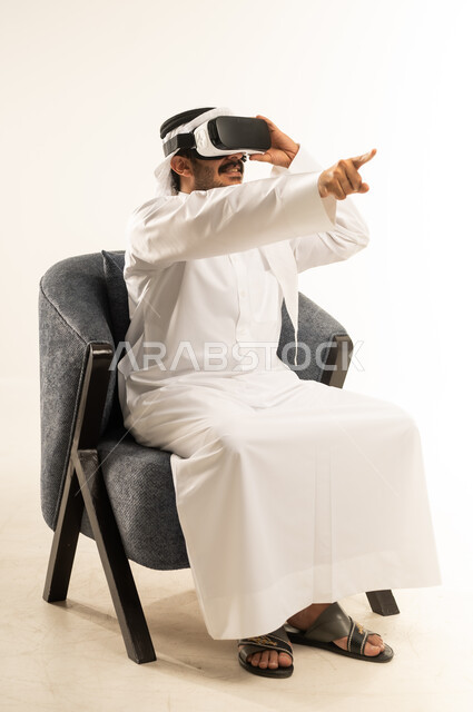 Trying to click on something, having fun watching through virtual reality glasses, portrait of a Qatari Gulf Arab man wearing traditional thobe and ghutra sitting on a comfortable sofa wearing 3D glasses raising his index finger up, pointing to a specific place with expressions of pleasure, white background