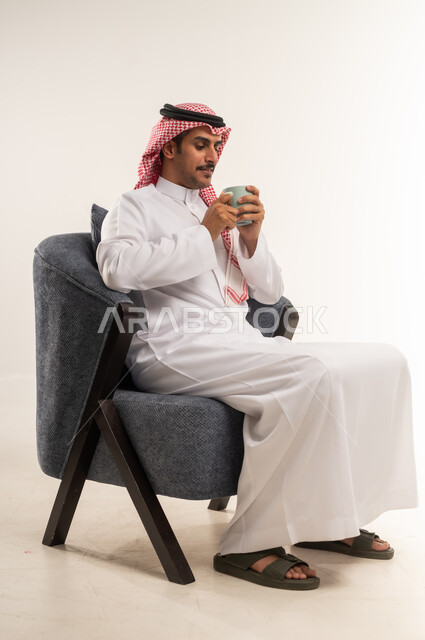 التلذذ بكوب من الشاي الساخن، محاولة الحصول على القليل من الراحة، الاسترخاء وتناول مشروبات دافئة، بورتريه من الجانب لشاب عربي خليجي بحريني يرتدي الثوب التقليدي والشماغ يجلس على كرسي ويمسك بيده فنجان من مشروبه المفضل، الطول الكامل للجسم، خلفية بيضاء