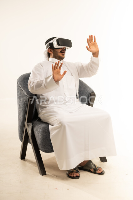 الاندماج والتفاعل ومحاكاة العالم الخيالي، بورتريه لرجل عربي خليجي قطري يرتدي الثوب التقليدي والغترة ونظارات الواقع الافتراضي يجلس على كرسي مريح يقوم بمد يديه الى الامام بتعابير صد شيء ما والدفاع عن النفس، دمج التسلية مع والتقنيات الحديثة، خلفية بيضاء