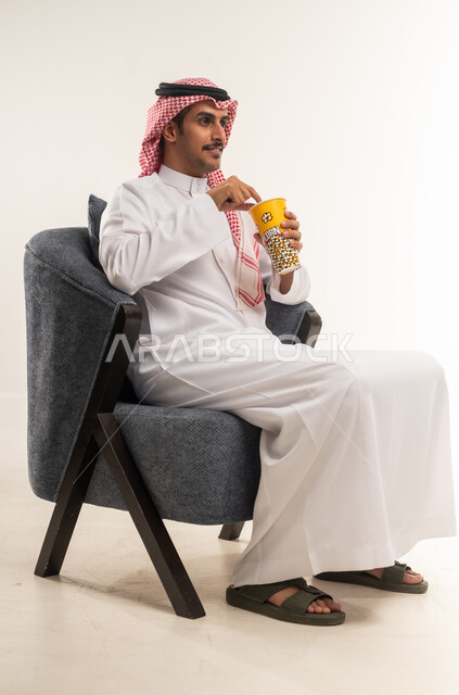 الجلوس ومشاهدة الأفلام في أوقات الفراغ، إيماءات الاندماج والتركيز مع الفيلم، الاستمتاع بتناول الفشار، بورتريه من الجانب لشاب عربي خليجي بحريني يرتدي الثوب التقليدي والشماغ يجلس على كرسي ويمسك بيده علبة من البوب كورن اللذيذ، الطول الكامل للجسم، خلفية بيضاء