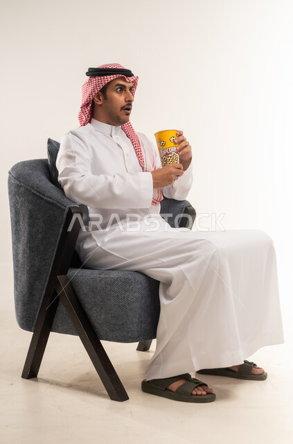 إيماءات الاندماج والتركيز مع الفيلم، الجلوس ومشاهدة الأفلام في أوقات الفراغ، الاستمتاع بتناول الفشار، بورتريه من الجانب لشاب عربي خليجي بحريني يرتدي الثوب التقليدي والشماغ يجلس على كرسي ويمسك بيده علبة من البوب كورن اللذيذ، الطول الكامل للجسم، خلفية بيضاء