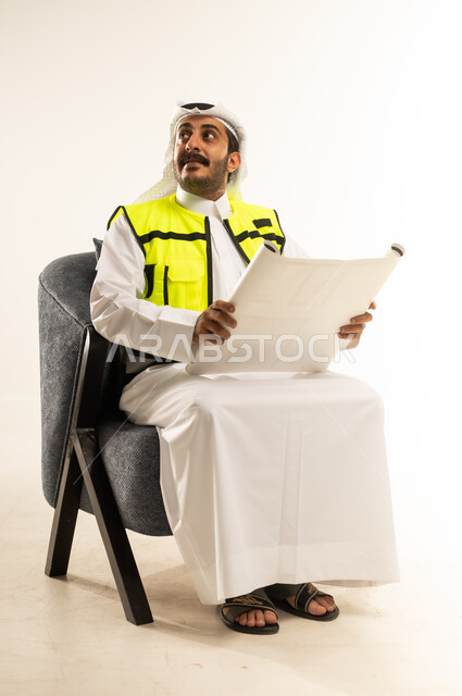 النظر الى مكان ما بتعابير السرور، إدارة ومتابعة المشروع, مفهوم الهندسة والانشاءات المعمارية, العمل في القطاع الهندسي، بورتريه لرجل عربي خليجي قطري يرتدي الثوب مع سترة الحماية والغترة يجلس على كرسي مريح يمسك مخططات الإعمار بيديه، خلفية بيضاء