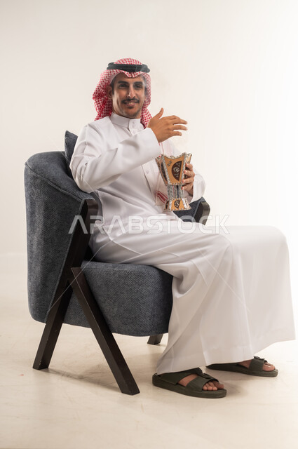 اتباع العادات والتقاليد العربية الأصيلة، التبخير واستخدام العطور والمطيبات، التبخر في الأعياد والمناسبات السعيدة، بورتريه من الجانب لشاب عربي خليجي بحريني يرتدي الثوب التقليدي والشماغ يجلس على كرسي ويمسك بيده المبخرة، الطول الكامل للجسم، خلفية بيضاء