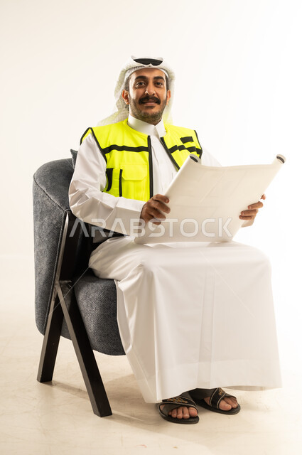 العمل في القطاع الهندسي، بورتريه لرجل عربي خليجي قطري يرتدي الثوب مع سترة الحماية والغترة يجلس على كرسي مريح يمسك مخططات الإعمار بيديه، النظر الى الكاميرا بتعابير الفخر والسرور، إدارة ومتابعة المشروع, مفهوم الهندسة والانشاءات المعمارية, خلفية بيضاء