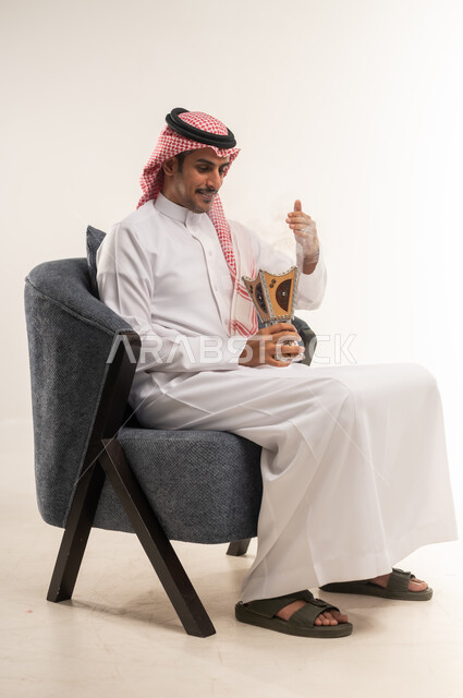التبخير واستخدام العطور والمطيبات، اتباع العادات والتقاليد العربية الأصيلة، التبخر في الأعياد والمناسبات السعيدة، بورتريه من الجانب لشاب عربي خليجي بحريني يرتدي الثوب التقليدي والشماغ يجلس على كرسي ويمسك بيده المبخرة، الطول الكامل للجسم، خلفية بيضاء