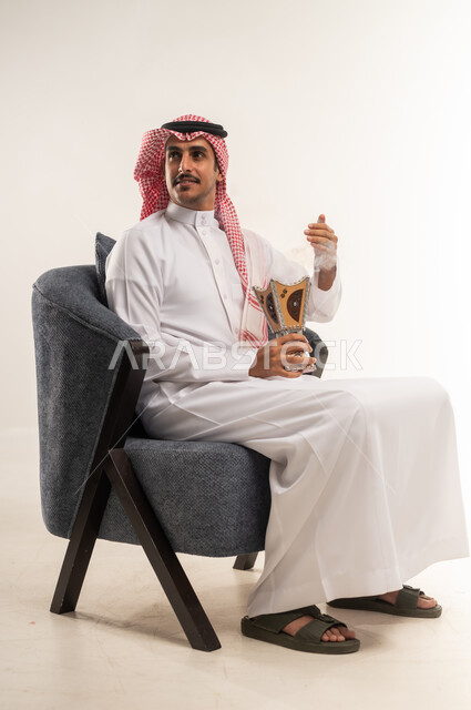التبخر في الأعياد والمناسبات السعيدة، اتباع العادات والتقاليد العربية الأصيلة، التبخير واستخدام العطور والمطيبات، بورتريه من الجانب لشاب عربي خليجي بحريني يرتدي الثوب التقليدي والشماغ يجلس على كرسي ويمسك بيده المبخرة، الطول الكامل للجسم، خلفية بيضاء