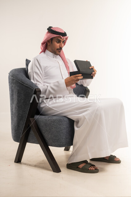 مفهوم المفاجأة والإهداء، إيماءات السعادة والدهشة بالحصول على هدية ثمينة، فن انتقاء الهدايا بعناية، بورتريه من الجانب لشاب عربي خليجي بحريني يرتدي الثوب التقليدي والشماغ يجلس على كرسي ويقوم بفتح العلبة، الطول الكامل للجسم، خلفية بيضاء