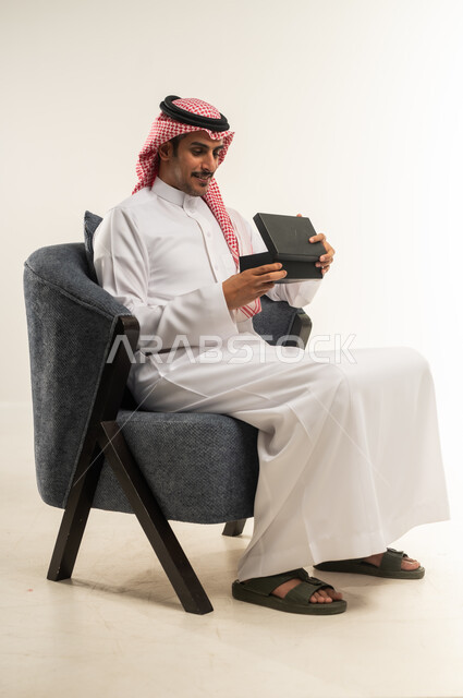 إيماءات السعادة والرضا بالحصول على هدية ثمينة، مفهوم المفاجأة والإهداء، فن انتقاء الهدايا بعناية، بورتريه من الجانب لشاب عربي خليجي بحريني يرتدي الثوب التقليدي والشماغ يجلس على كرسي ويقوم بفتح العلبة، الطول الكامل للجسم، خلفية بيضاء