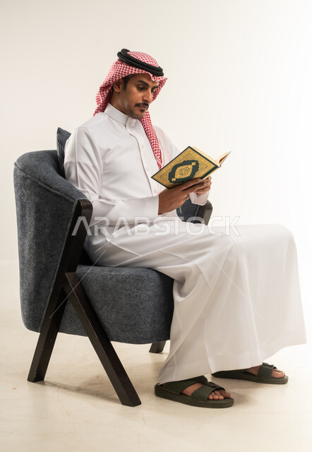 تلاوة وترتيل كتاب الله، حفظ وتفسير كلام الله عز وجل، أداء العبادات والتقرب إلى الله تعالى، التعبد والتضرع بخشوع، بورتريه من الجانب لشاب عربي خليجي بحريني يرتدي الثوب التقليدي والشماغ يجلس على كرسي ويمسك القرآن الكريم بيديه، الطول الكامل للجسم، خلفية بيضاء