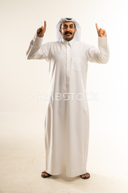 حركات يدين واشارات تدل على هذا المكان (هنا)، تحديد موقع شيء معين، بورتريه لرجل عربي خليجي قطري يرتدي الثوب التقليدي والغتره يشير باصبع السبابة إلى الأعلى، مفهوم الأناقة والاهتمام بالمظهر الخارجي، الجسم بالطول الكامل، خلفيه بيضاء