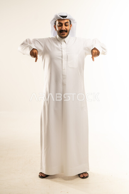 تأكيد حدوث شيء هذه اللحظة, حركات تدل على هذا المكان (هنا)، تحديد موقع معين، بورتريه لرجل عربي خليجي قطري يرتدي الثوب التقليدي والغترة يشير بإصابع السبابة نحو الأسفل, النظر إلى الكاميرا بايماءات السرور، الطول الكامل للجسم, خلفية بيضاء