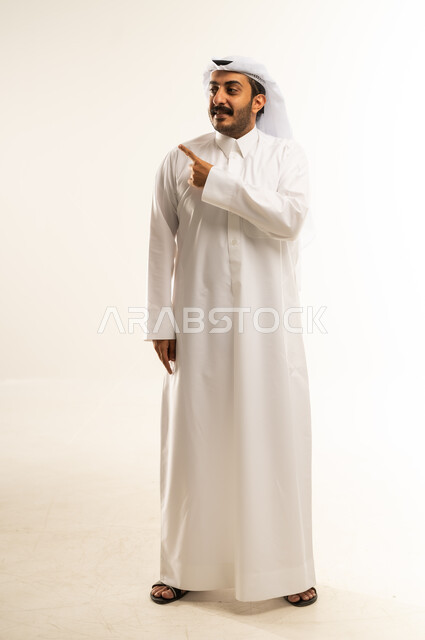 تحديد موقع شيء معين، إشارات وحركات يدين تدل على هذا المكان (هنا)، بورتريه لرجل عربي خليجي قطري يرتدي الثوب التقليدي والغتره يشير باصبع السبابة إلى الأعلى، مفهوم الأناقة والاهتمام بالمظهر الخارجي، الجسم بالطول الكامل، خلفيه بيضاء