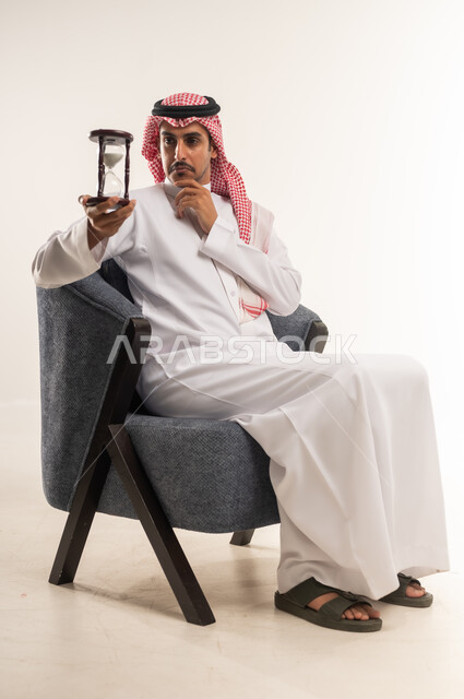 التخطيط لتحقيق الأهداف في المستقبل، إدراك قيمة اللحظات الثمينة في الحياة، مراقبة مرور الوقت، انتظار شيء ما بفارغ الصبر، بورتريه من الجانب لشاب عربي خليجي بحريني يرتدي الثوب التقليدي والشماغ يجلس على كرسي ويمسك الساعة الرملية، الطول الكامل للجسم، خلفية بيضاء