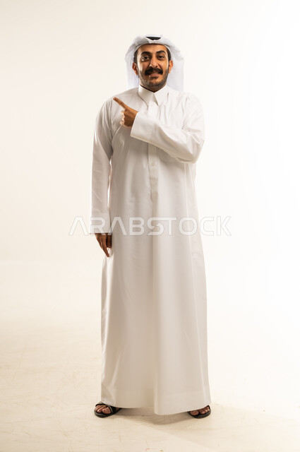 تحديد موقع شيء معين، إشارات وحركات يدين تدل على هذا المكان (هنا)، بورتريه لرجل عربي خليجي قطري يرتدي الثوب التقليدي والغتره يشير باصبع السبابة إلى الأعلى، مفهوم الأناقة والاهتمام بالمظهر الخارجي، الجسم بالطول الكامل، خلفيه بيضاء