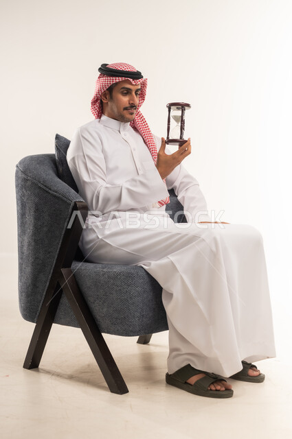 مراقبة مرور الوقت، إدراك قيمة اللحظات الثمينة في الحياة، التخطيط لتحقيق الأهداف في المستقبل، انتظار شيء ما بفارغ الصبر، بورتريه من الجانب لشاب عربي خليجي بحريني يرتدي الثوب التقليدي والشماغ يجلس على كرسي ويمسك الساعة الرملية، الطول الكامل للجسم، خلفية بيضاء