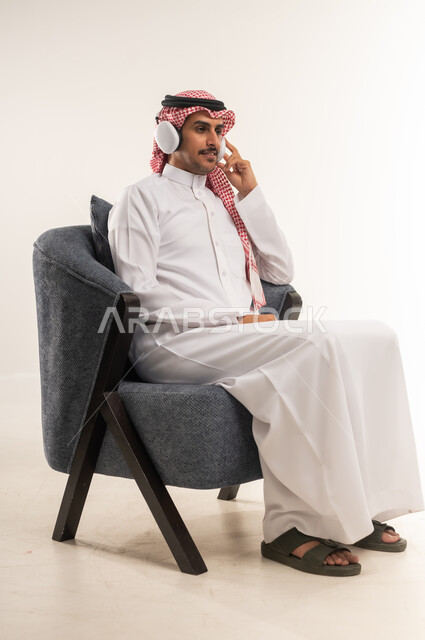 استخدام تقنية الهيدفون، الاستماع إلى الموسيقا والبودكاست، دمج التقنيات والأجهزة الحديثة والتكنولوجيا بالحياة اليومية، بورتريه من الجانب لشاب عربي خليجي بحريني يرتدي الثوب التقليدي والشماغ يجلس على كرسي مريح ويضع سماعة الأذنين، الطول الكامل للجسم، خلفية بيضاء