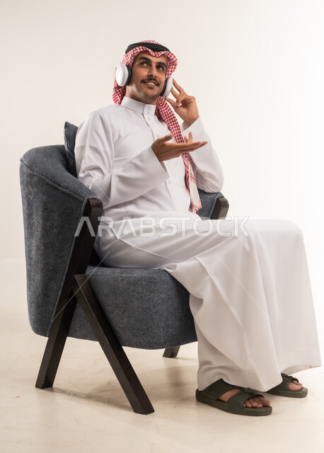 دمج التقنيات والأجهزة الحديثة والتكنولوجيا بالحياة اليومية، الاستماع إلى الموسيقا والبودكاست، استخدام تقنية الهيدفون، بورتريه من الجانب لشاب عربي خليجي بحريني يرتدي الثوب التقليدي والشماغ يجلس على كرسي مريح ويضع سماعة الأذنين، الطول الكامل للجسم، خلفية بيضاء