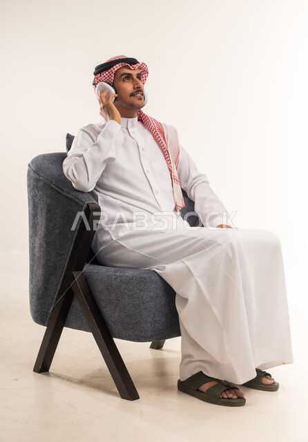 الاستماع إلى الموسيقا والبودكاست، استخدام تقنية الهيدفون، دمج التقنيات والأجهزة الحديثة والتكنولوجيا بالحياة اليومية، بورتريه من الجانب لشاب عربي خليجي بحريني يرتدي الثوب التقليدي والشماغ يجلس على كرسي مريح ويضع سماعة الأذنين، الطول الكامل للجسم، خلفية بيضاء