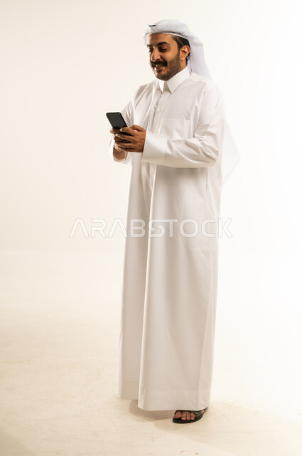 دمج التكنولوجيا في الحياة اليومية، استخدام التقنيات والأجهزة الحديثة، تصفح التطبيقات ومواقع التواصل الاجتماعي عبر الانترنت، دردشة ومحادثات وتعارف، بورتريه لرجل عربي خليجي قطري يرتدي الثوب التقليدي يقوم باستخدام الهاتف المحمول، الجسم بالطول الكامل، خلفية بيضاء