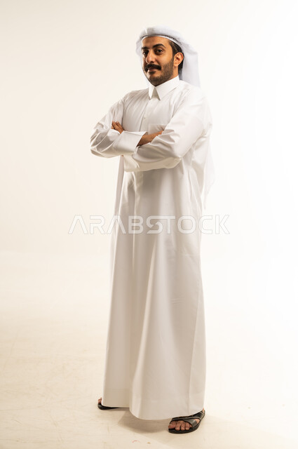 الاهتمام بالأناقة والمظهر الخارجي، النظر إلى الكاميرا بايماءات الجدية والسرور، مفهوم الرجولة والقوة والثقة بالنفس، بورتريه لرجل عربي خليجي قطري يرتدي الثوب التقليدي والغترة يقف بأيدي متقاطعة بتعابير الحزم، الطول الكامل للجسم، خلفية بيضاء