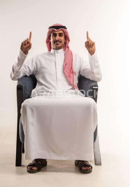 اشارة رقم واحد، شاب سعودي ينظر إلى الكاميرا بإيماءات الفرح والسرور، بورتريه  لرجل عربي خليجي بحريني مبتسم يرتدي الثوب التقليدي والشماغ يجلس على كرسي مريح يشير بإصبعه السبابة للاعلى  إلى شيء محدد بتعابير هذا المكان هنا، خلفية بيضاء .