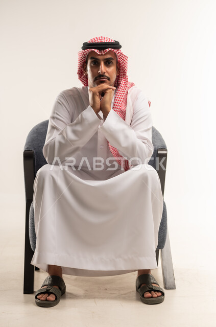 الشعور بالحيرة وعدم التأكد، إيماءات التأمل والتمعن والتركيز, بورتريه لرجل عربي خليجي بحريني يرتدي الثوب التقليدي والشماغ يجلس على كرسي يضع يديه خلف ذقنه، شاب سعودي ينظر الى الكاميرا بتعابير التفكير والشرود، التخطيط لحل مشكلة ما، خلفية بيضاء