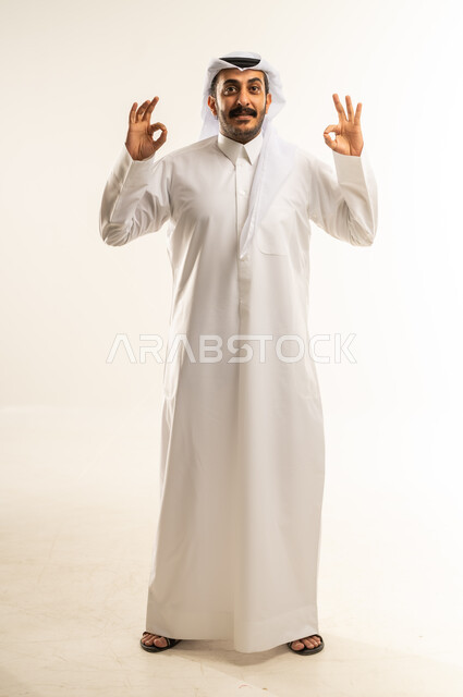 التعبير عن الإعجاب والتقدير، رفع اليد بعلامة الجودة والقبول، الامتياز والعلامة الكاملة، الشعور بالرضا والاستحسان، بورتريه لرجل عربي خليجي قطري مبتسم يرتدي الثوب التقليدي والغتره ينظر إلى الكاميرا بإيماءات السعادة والسرور، الجسم بالطول الكامل، خلفية بيضاء