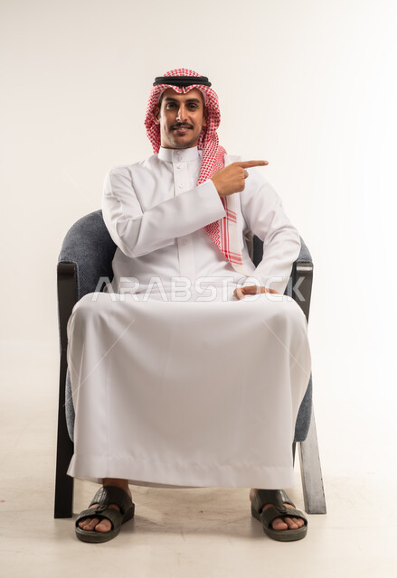 الاشارة والتحديد لعنصر معين، شاب سعودي ينظر إلى الكاميرا بإيماءات الفرح والسرور، بورتريه لشاب عربي خليجي بحريني مبتسم يرتدي الثوب التقليدي والشماغ يجلس على كرسي مريح يشير بإصبعه السبابة جانبا  إلى شيء محدد بتعابير هذا المكان هنا، خلفية بيضاء .