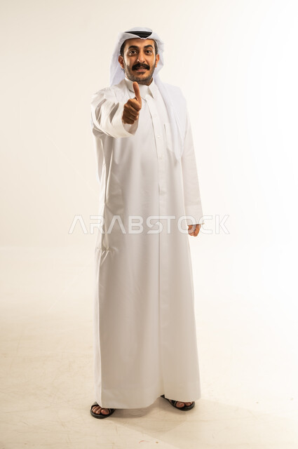 علامة الجودة والامتياز, بورتريه لرجل عربي خليجي قطري مبتسم يرتدي الثوب التقليدي والغترة يرفع ابهامه بإيماءات الموافقة والقبول, الاعجاب والرضا لتحقيق أمر معين, صورة الجسم الكامل, خلفية بيضاء