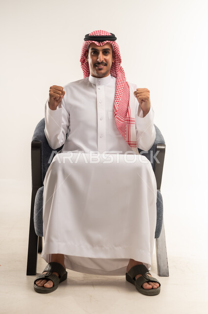 مفهوم الانجاز وتحقيق الاهداف، بورتريه لرجل عربي خليجي بحريني مبتسم يرتدي الثوب التقليدي والشماغ يجلس على كرسي مريح يرفع قبضة يديه للاعلى بإيماءات الفوز والانتصار والنجاح, شاب سعودي ينظر الى الكاميرا بتعابير السعادة والقوة والسرور، خلفية بيضاء
