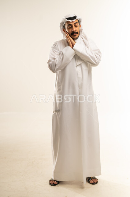 النعاس والحاجه الى النوم والراحة، بورتريه لرجل عربي خليجي قطري يرتدي الثوب التقليدي والغترة يضع يده تحت رأسه بإيماءات وجه ويد تدل على النعاس, الإحساس بالتعب الكسل والخمول، السهر لأوقات متأخرة، صوره الجسم بالطول الكامل، خلفيه بيضاء
