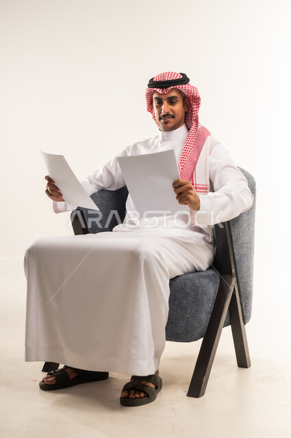 قراءة الملاحظات والمعلومات، بورتريه لرجل عربي خليجي بحريني يرتدي الثوب التقليدي والشماغ يجلس على كرسي مريح يقوم بقراءة وتدقيق الاوراق، شاب سعودي ينظر الى الملفات بتعابير التركيز والتمعن، مفهوم انجاز العمل، متابعة الخطط والمشاريع، خلفية بيضاء