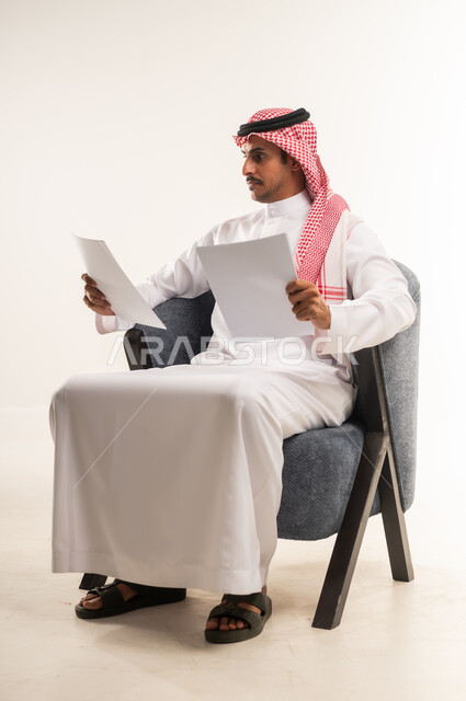 قراءة الملاحظات والمعلومات، بورتريه لرجل عربي خليجي بحريني يرتدي الثوب التقليدي والشماغ يجلس على كرسي مريح يقوم بقراءة وتدقيق الاوراق، شاب سعودي ينظر الى الملفات بتعابير التركيز والتمعن، مفهوم انجاز العمل، متابعة الخطط والمشاريع، خلفية بيضاء