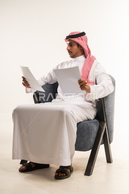 مفهوم انجاز العمل، متابعة الخطط والمشاريع، قراءة الملاحظات والمعلومات، بورتريه لرجل عربي خليجي بحريني يرتدي الثوب التقليدي والشماغ يجلس على كرسي مريح يقوم بقراءة وتدقيق الاوراق، شاب سعودي ينظر الى الملفات بتعابير التركيز والتمعن، خلفية بيضاء