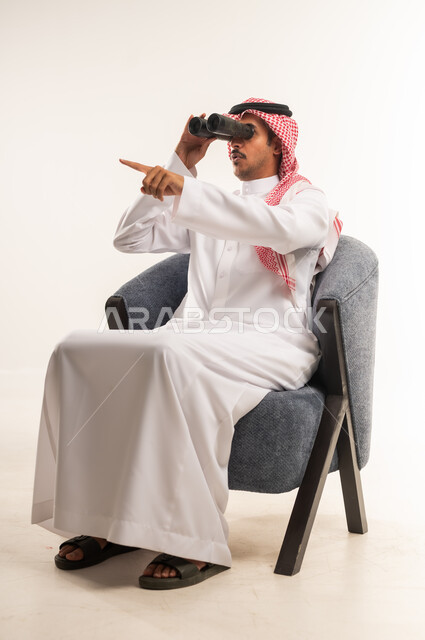رؤية الاشياء البعيدة عبر المنظار، حب المغامرة والاستكشاف، بورتريه لرجل عربي خليجي بحريني يرتدي الثوب التقليدي والشماغ يجلس على كرسي مريح ينظر من خلال المنظار، شاب سعودي يشير باصبع السبابة لمكان ما, التخطيط المستقبلي للأهداف والطموحات، خلفية بيضاء