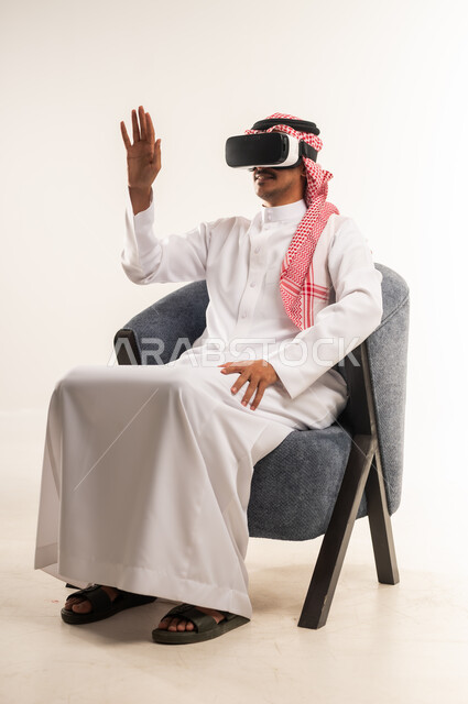 الاندماج والتفاعل ومحاكاة العالم الخيالي, بورتريه لرجل عربي خليجي بحريني يرتدي الثوب التقليدي والشماغ يجلس على كرسي مريح يضع نظارات الواقع الافتراضي يقوم برفع وفتح كف يده للاعلى بتعابير التوقف عن فعل امر ما, شاب سعودي يستخدم نظارة الفي آر، خلفية بيضاء